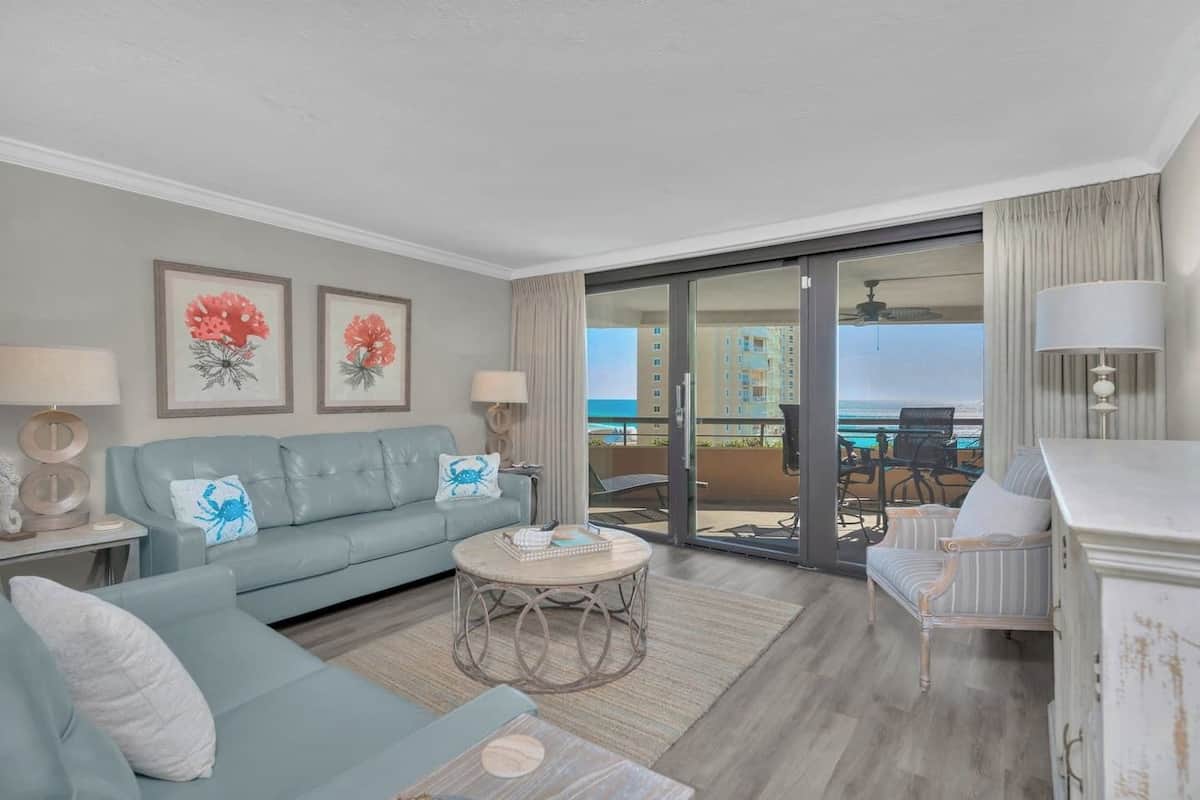 A modern living room with a light blue sofa, a coffee table, and a view of the ocean through large sliding doors.