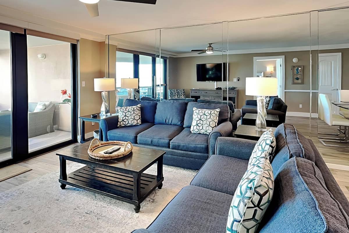 A modern living room with a large sectional sofa, a coffee table, and a TV. There is a sliding glass door leading to an outdoor area.