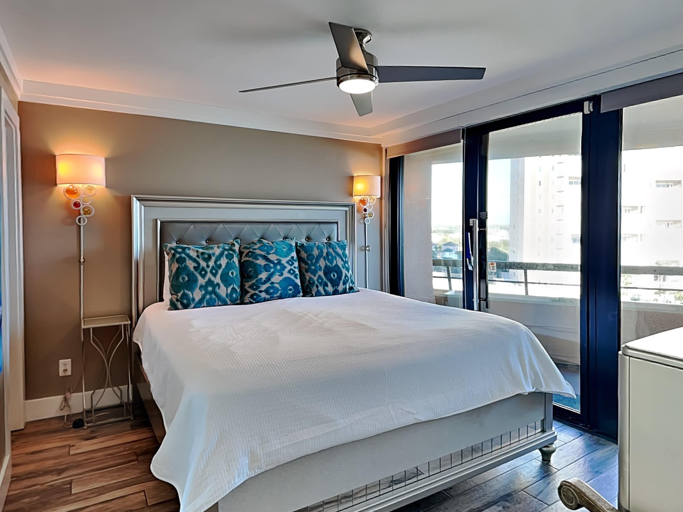 A hotel room with a bed, ceiling fan, two bedside lamps, and a view of the city through large windows.