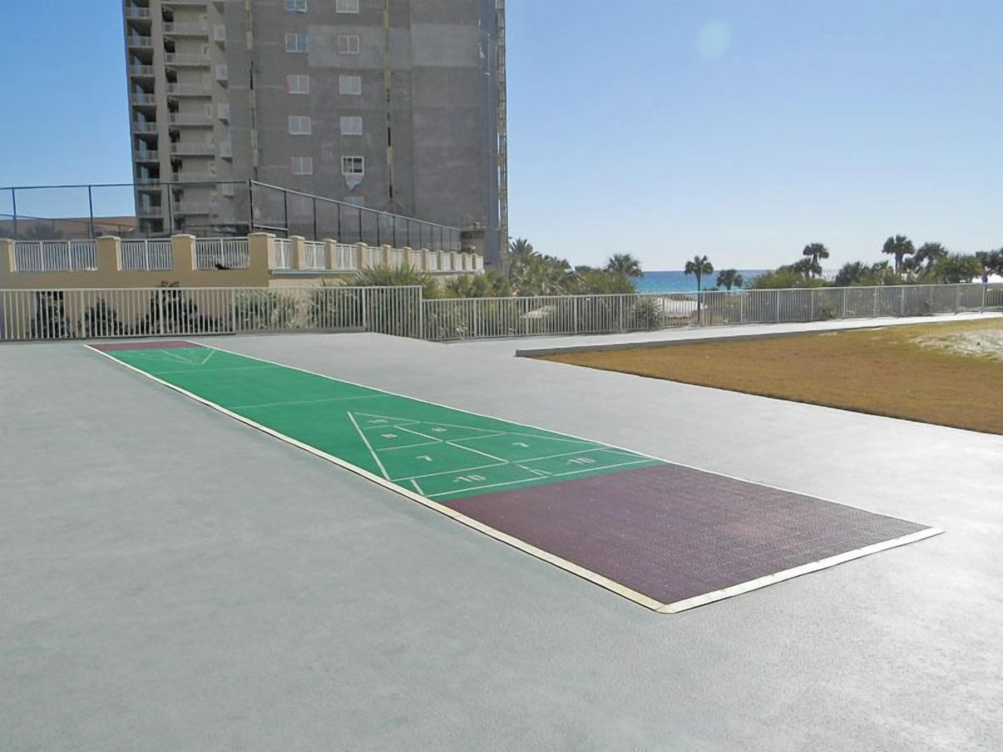 A shuffleboard court with a green playing surface and a purple end zone, surrounded by a concrete area and a metal fence.