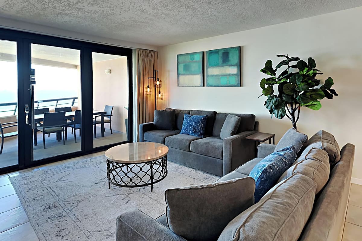 A modern living room with a grey sofa set, a coffee table, and a large plant. There is a sliding glass door leading to an outdoor seating area.