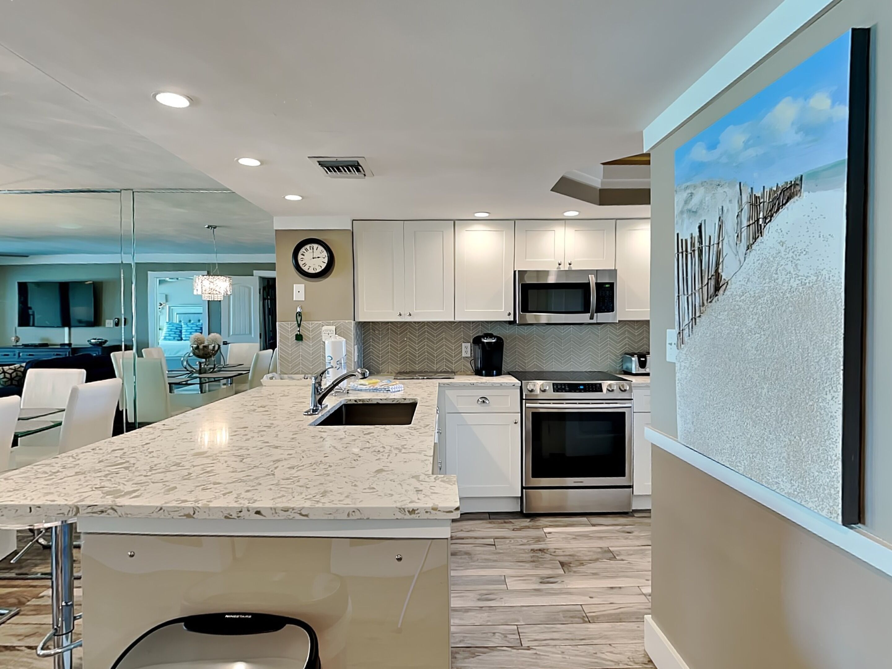 A modern kitchen with white cabinets, a large island with a sink, a microwave, and an oven. There is a clock on the wall and a painting of a beach scene.