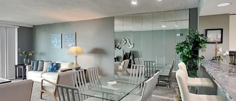 Uma sala de estar moderna com uma mesa de jantar de vidro, cadeiras estofadas cinzas, um sofá com almofadas estampadas, uma mesinha de apoio com uma luminária e um espelho grande.