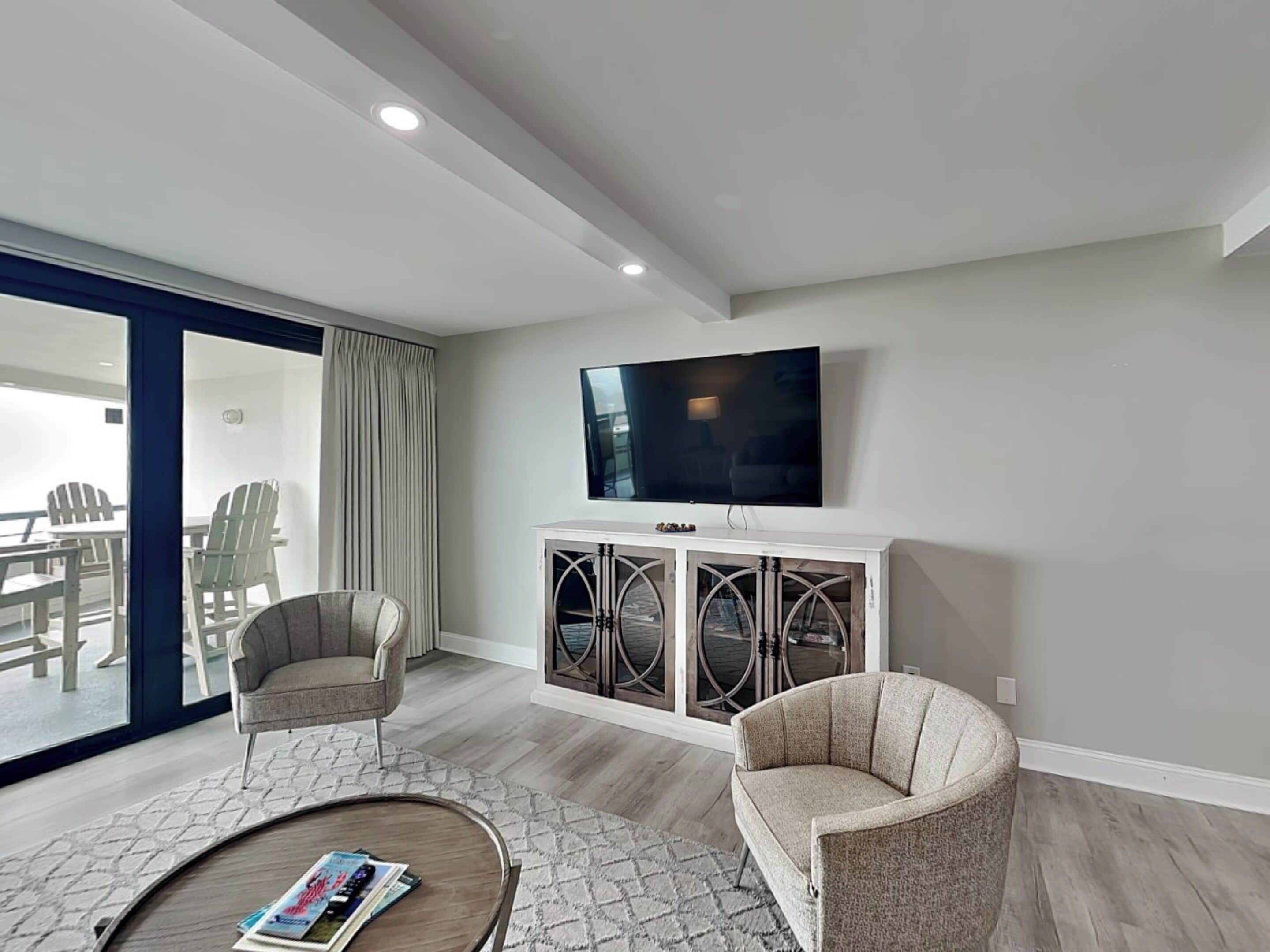 A modern living room with a flat-screen TV, two armchairs, a coffee table, and a glass door leading to an outdoor area.