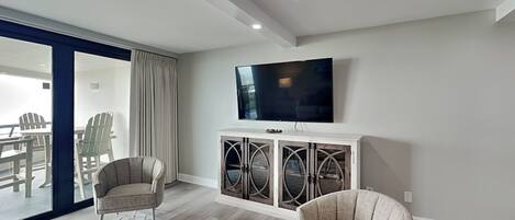 A modern living room with a flat-screen TV, two armchairs, a coffee table, and a glass door leading to an outdoor area.