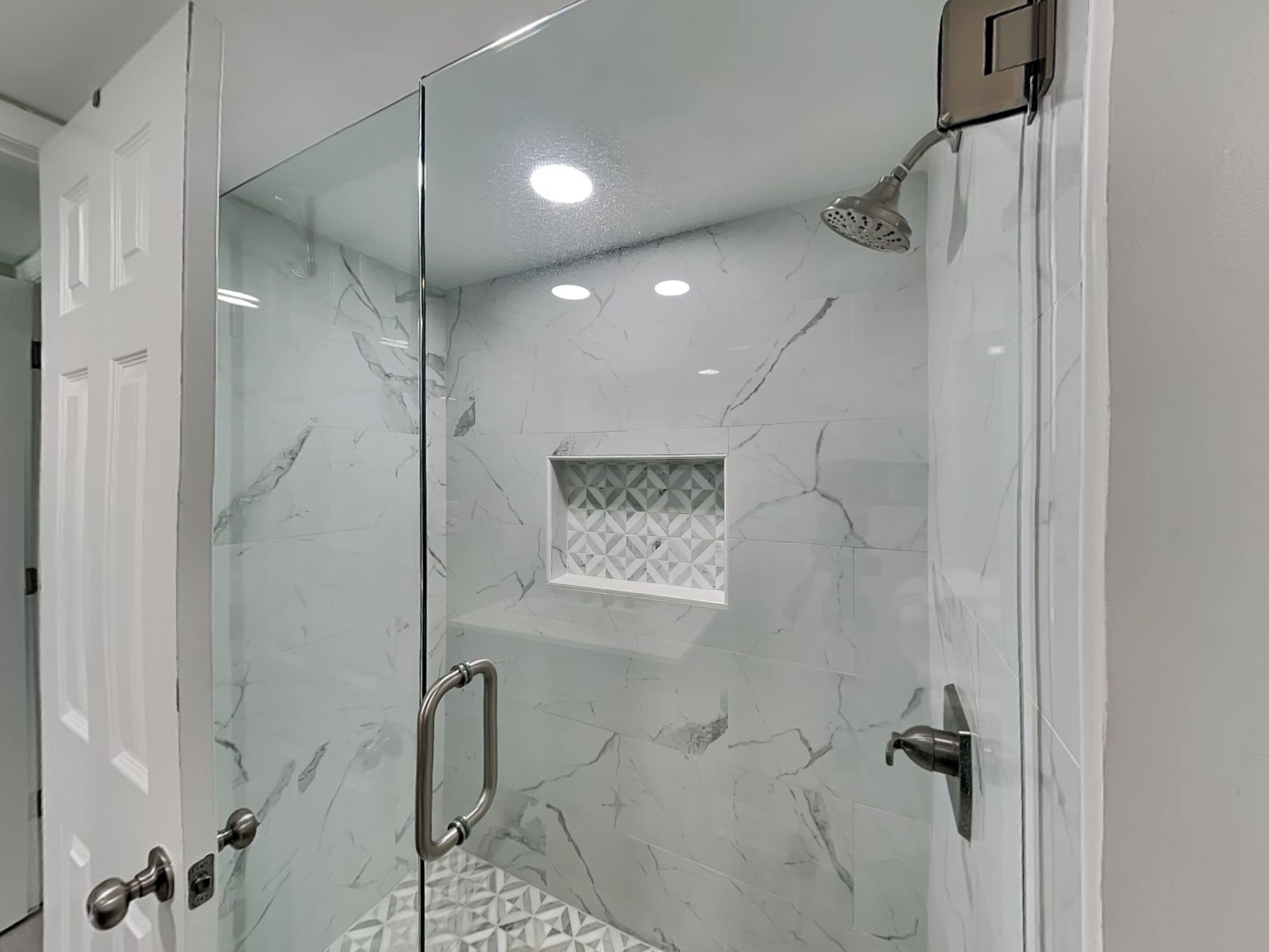 A glass-enclosed shower with a handheld showerhead, a geometric-patterned tile wall, and a recessed ceiling light.