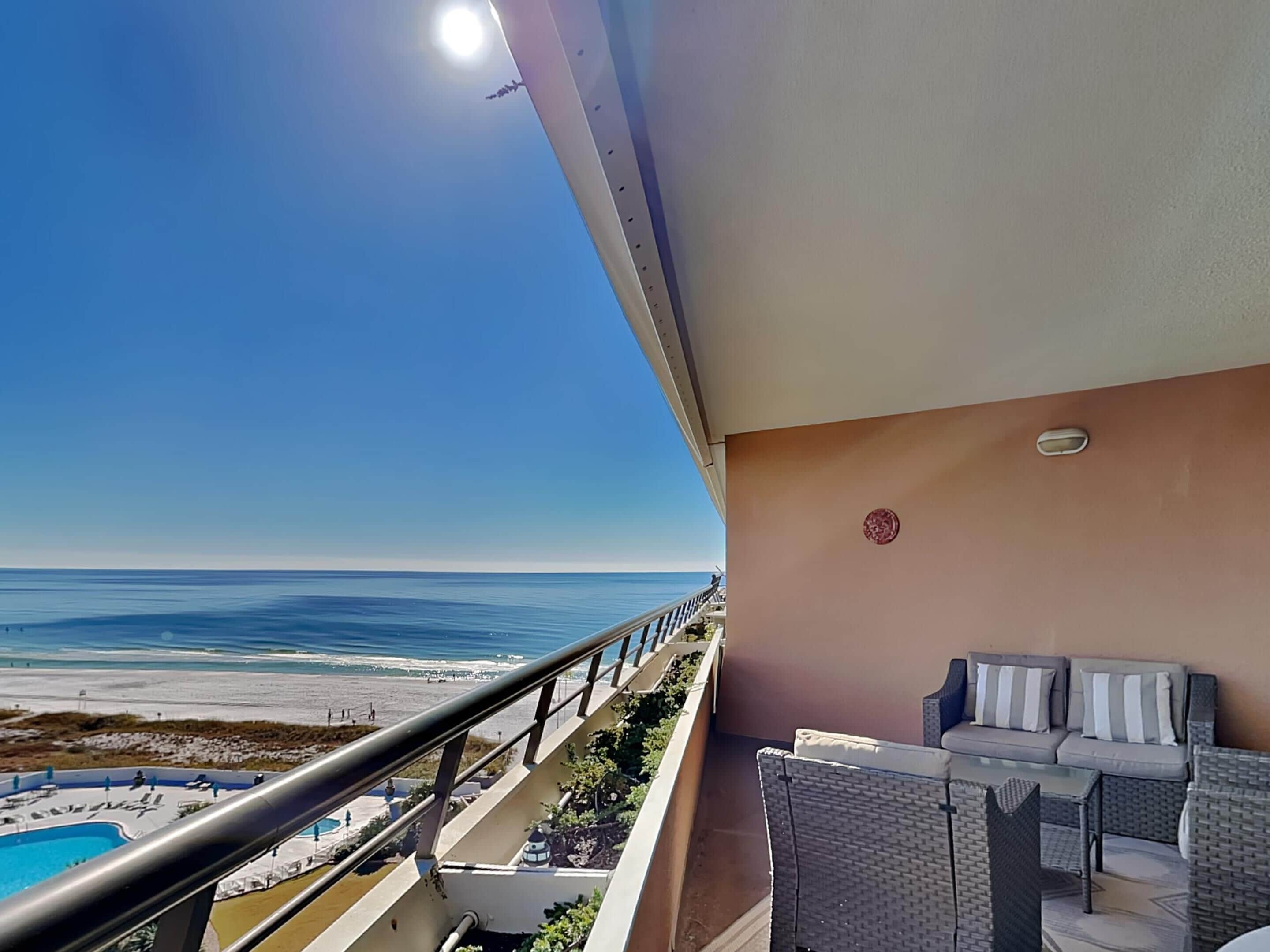 A balcony with a view of the beach and ocean, a pool, and outdoor furniture.