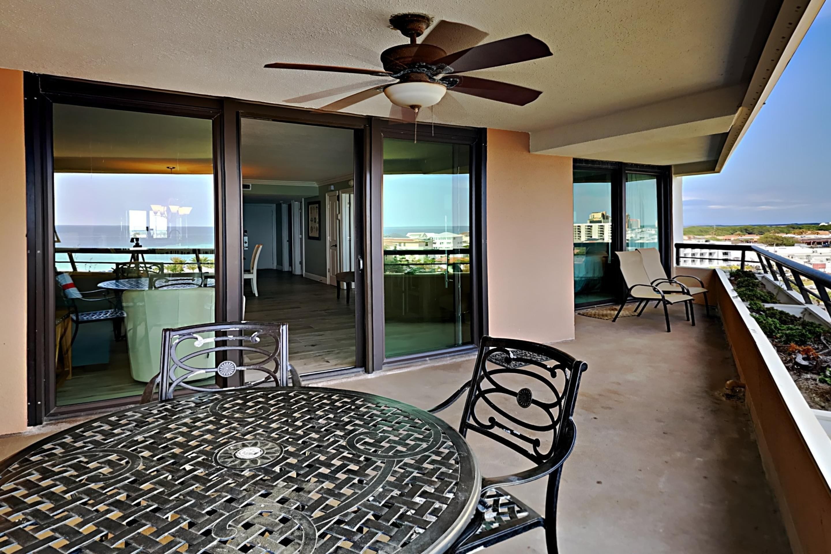 Ein Balkon mit Tisch und Stühlen, einem Deckenventilator und Blick auf das Meer und Gebäude.