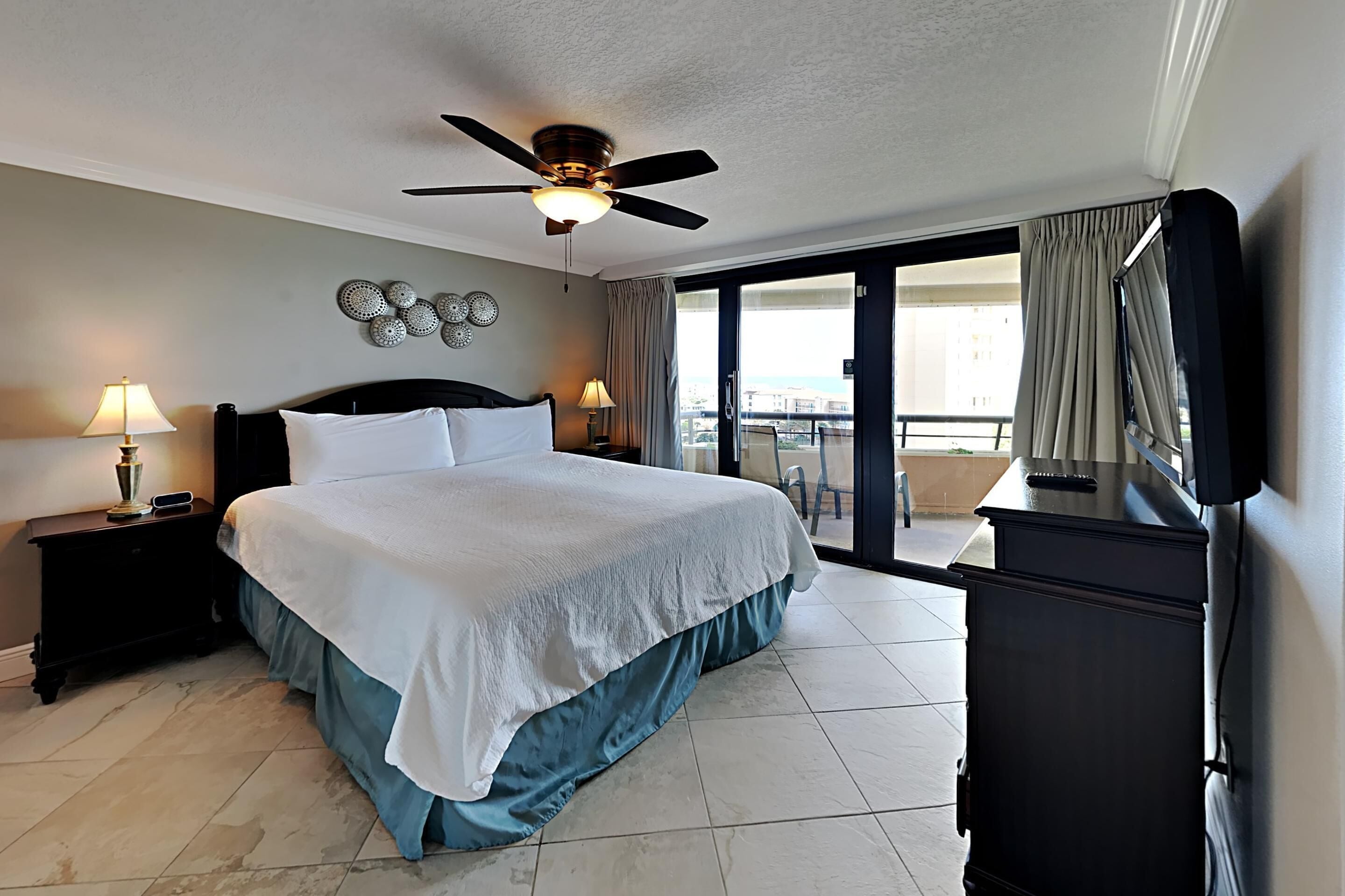 A hotel room with a large bed, two bedside tables with lamps, a ceiling fan, and a view of the outside balcony.