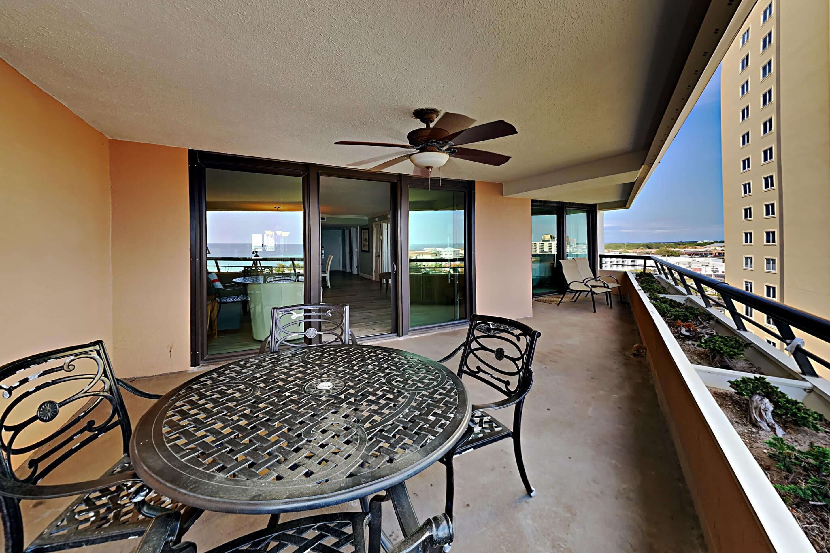 A balcony with a round metal table and chairs, a ceiling fan, and a view of the city and ocean.