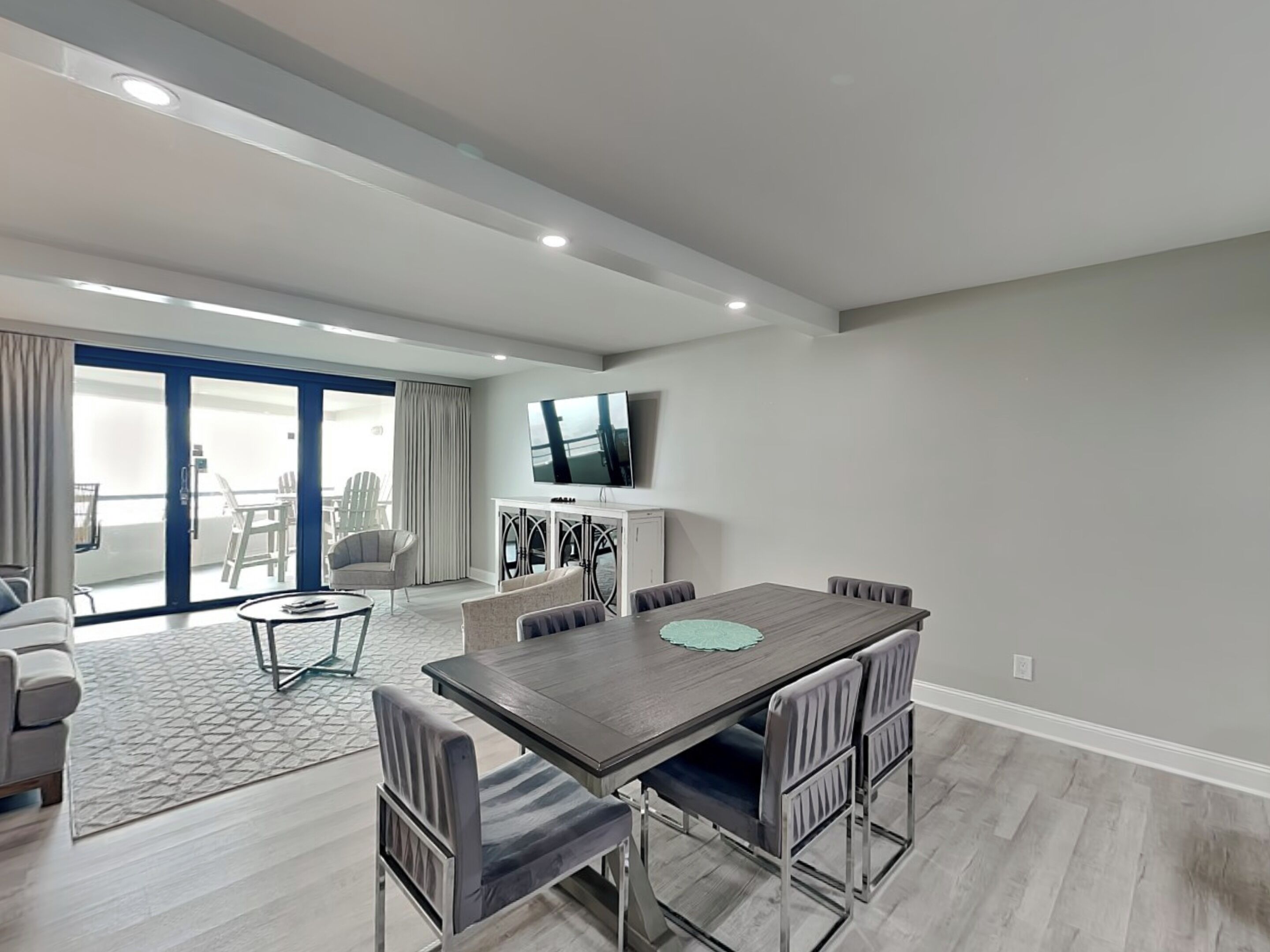 A modern living room with a dining area, a flat-screen TV mounted on the wall, and a balcony with outdoor seating.
