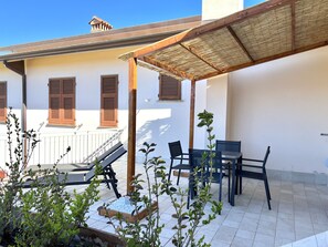 A covered outdoor seating area with a table and chairs, a wooden pergola, and a white building with brown shutters in the background. - Abitatt (Fosdinovo)