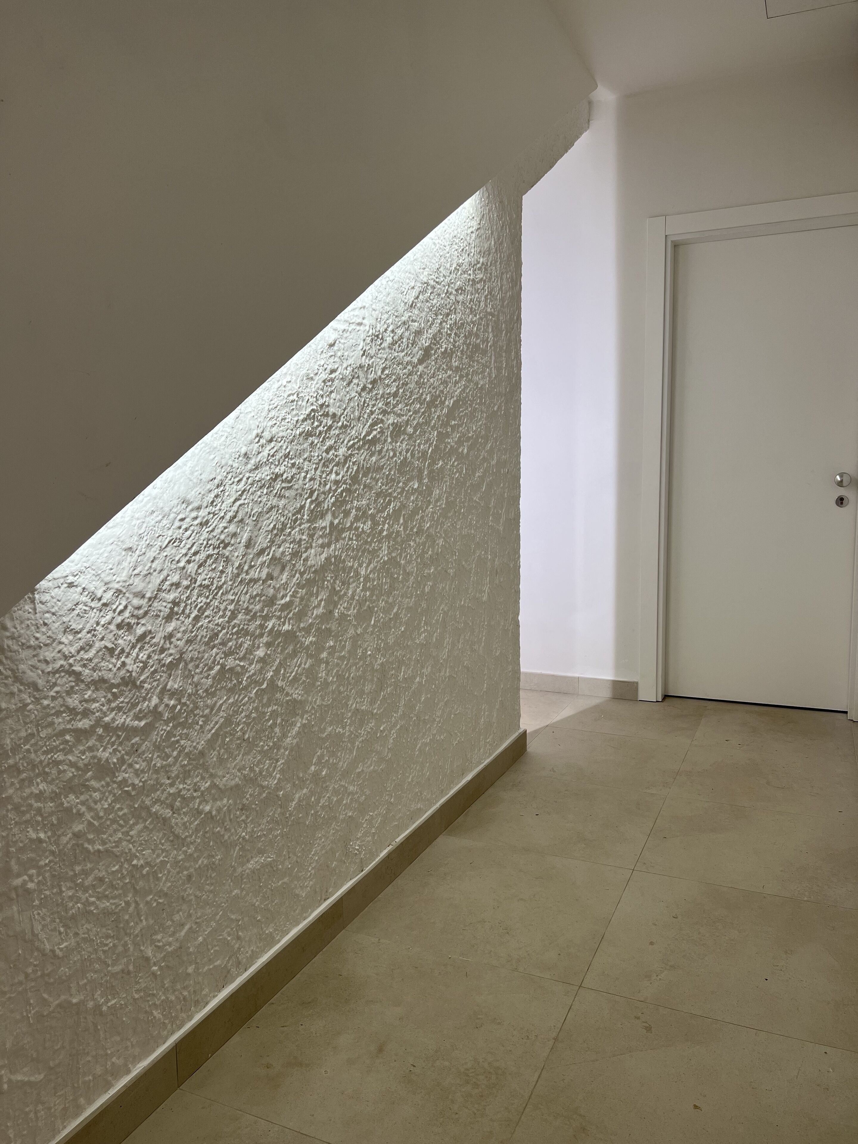 Un couloir avec un mur blanc à texture, une porte et un sol carrelé.