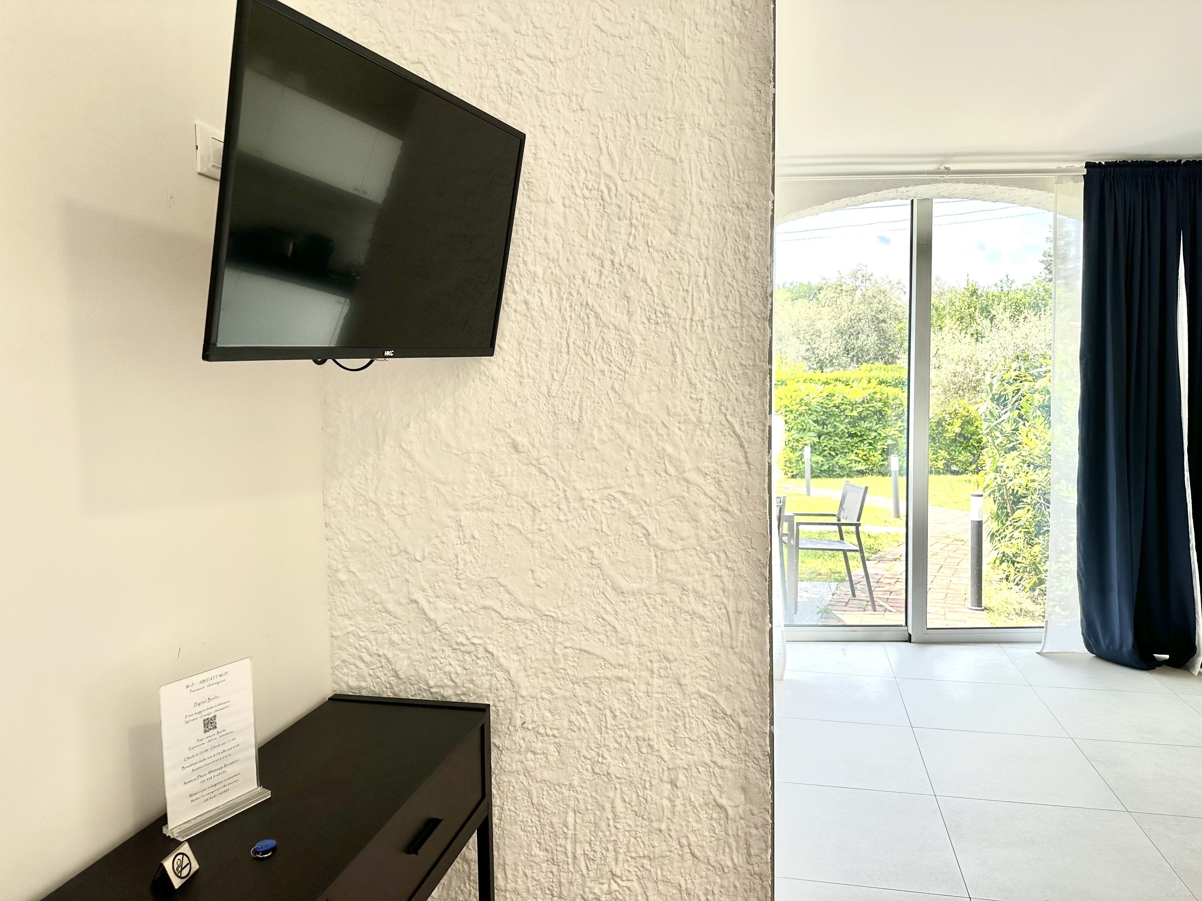Habitación con una televisión de pantalla plana montada en la pared, una mesita con un cartel y vistas a un balcón con una silla y plantas.