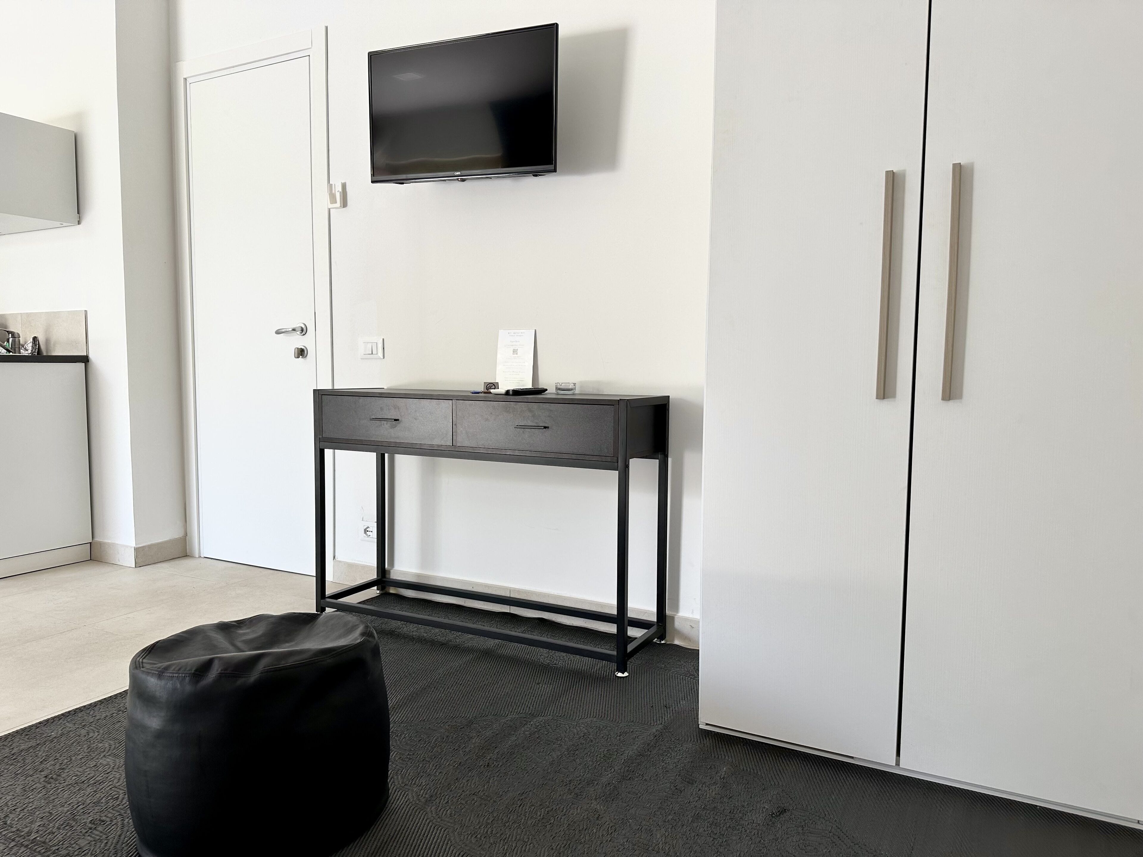 Une chambre moderne dotée d’un téléviseur à écran plat fixé au mur, d’un bureau avec deux tiroirs, d’un pouf noir et d’une armoire à deux portes.