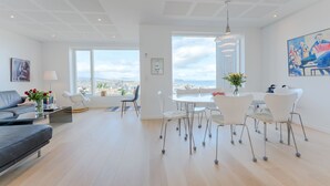 A modern living room with a sofa, dining table, and chairs, large windows with a city view, and a ceiling with a unique pattern. - Sea & City View - Luxurious - Hiking (Argir)