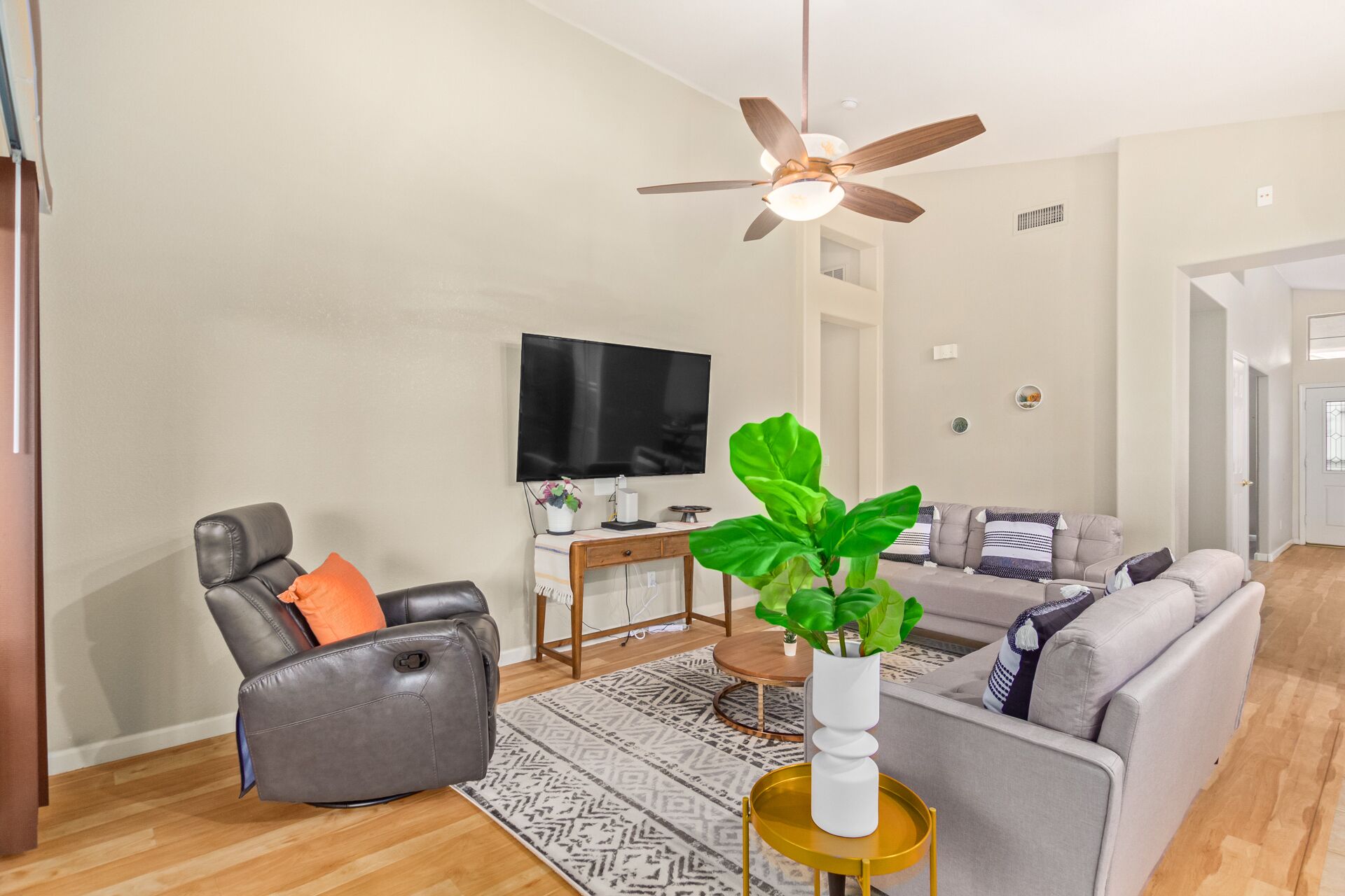 Ein modernes Wohnzimmer mit einem Ledersessel, einem grauen Sofa, einem Couchtisch, einem Fernseher und einem Deckenventilator.