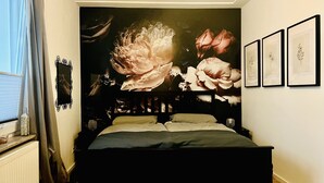 A bedroom with a large floral tapestry, a bed with two pillows, a bedside table, and a window with curtains. - Schöne Ruhige Zentrale Designerwohnung (Hannover)