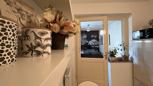 A narrow hallway with a door leading to a bedroom. On the left, there are decorative items including a leopard-print mug and a vase with flowers. A radiator is visible on the wall, and a microwave is mounted above the counter on the right. - Schöne Ruhige Zentrale Designerwohnung (Hannover)