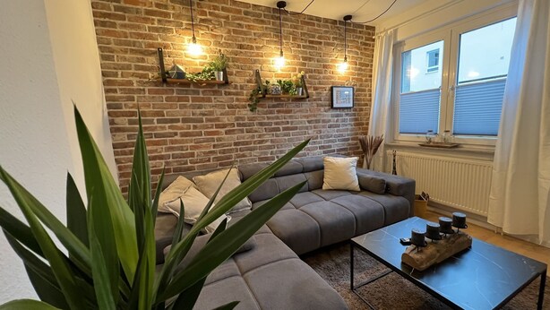 A living room with a brick wall, a grey sofa, a coffee table, and a window with blinds. - Schöne Ruhige Zentrale Designerwohnung (Hannover)
