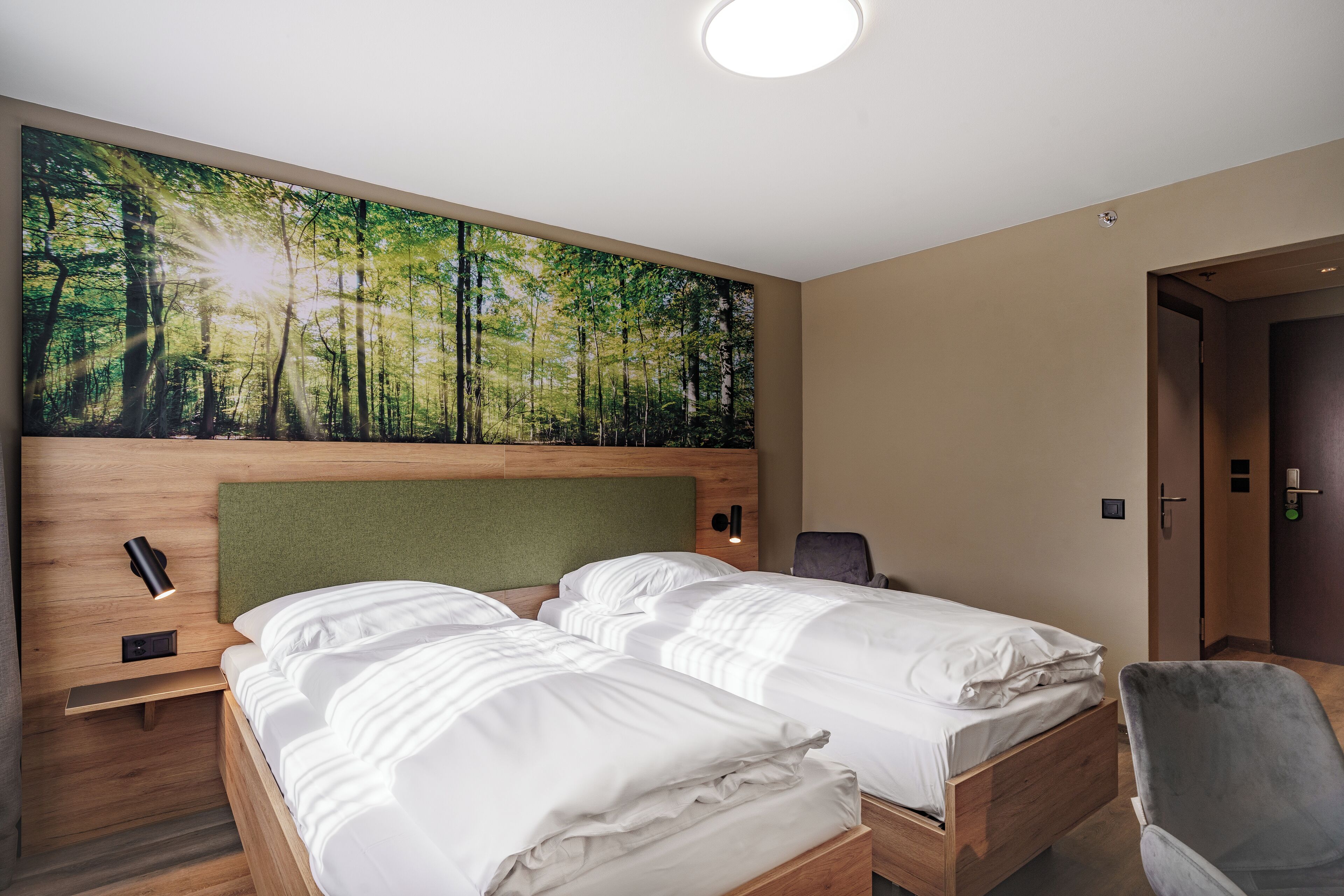 Ein Hotelzimmer mit einer großen Wandmalerei zum Thema Wald, zwei Betten mit weißer Bettwäsche, einem Holzkopfende, einem Nachttisch mit Lampe und einem Sessel.
