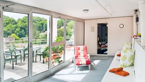 A balcony with a view of a green hillside, a seating area with chairs and a table, and a kitchen area with a clock and a plant. - Hotel Linde 24h Self Check-in (Baden)