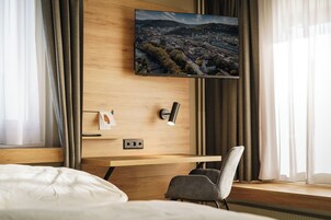Quarto de hotel com cama, escrivaninha com cadeira, televisão de tela plana na parede e janela com cortinas.