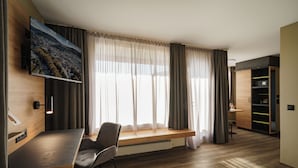 A modern room with a wooden desk, a chair, a TV mounted on the wall, a window with sheer curtains, and a built-in shelving unit. - Hotel Linde 24h Self Check-in (Baden)