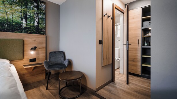 A modern hotel room with a bed, a small round table, a chair, a wardrobe, and a large forest-themed wall mural. - Hotel Linde 24h Self Check-in (Baden)