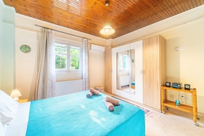 A bedroom with a bed, curtains, a window, a mirror, a wooden shelf with speakers, and a radiator. - Holiday Home "Corfu House in River" with Garden & Wi-Fi (Kerkyra)