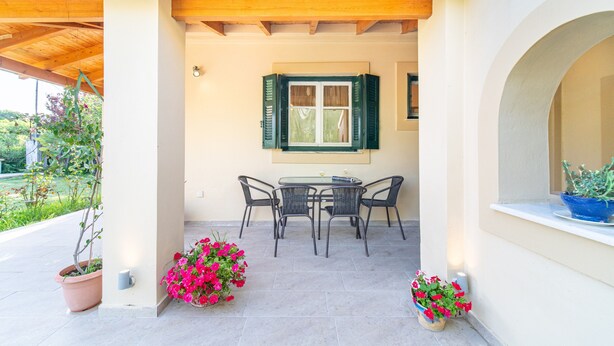 Une terrasse couverte avec une table et des chaises, des plantes en pot et une fenêtre avec des volets.