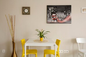 Un coin repas avec une table blanche, des chaises jaunes, une plante en pot, une horloge murale et un tableau représentant un paysage urbain.