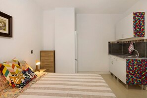 Une chambre compacte comprenant un lit, une table de chevet, une lampe et une commode. À côté se trouve une petite cuisine équipée d’un évier et de placards.
