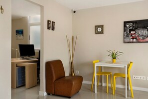 A modern living room with a brown sofa, a dining table with yellow chairs, a white desk with a computer, and a vase with sticks.