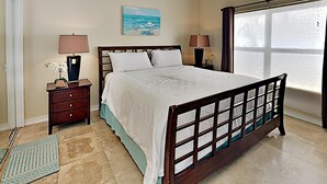 Un dormitorio con una cama de madera, dos mesitas de noche con lámparas, un ventilador de techo, un cuadro en la pared y una ventana con persianas.