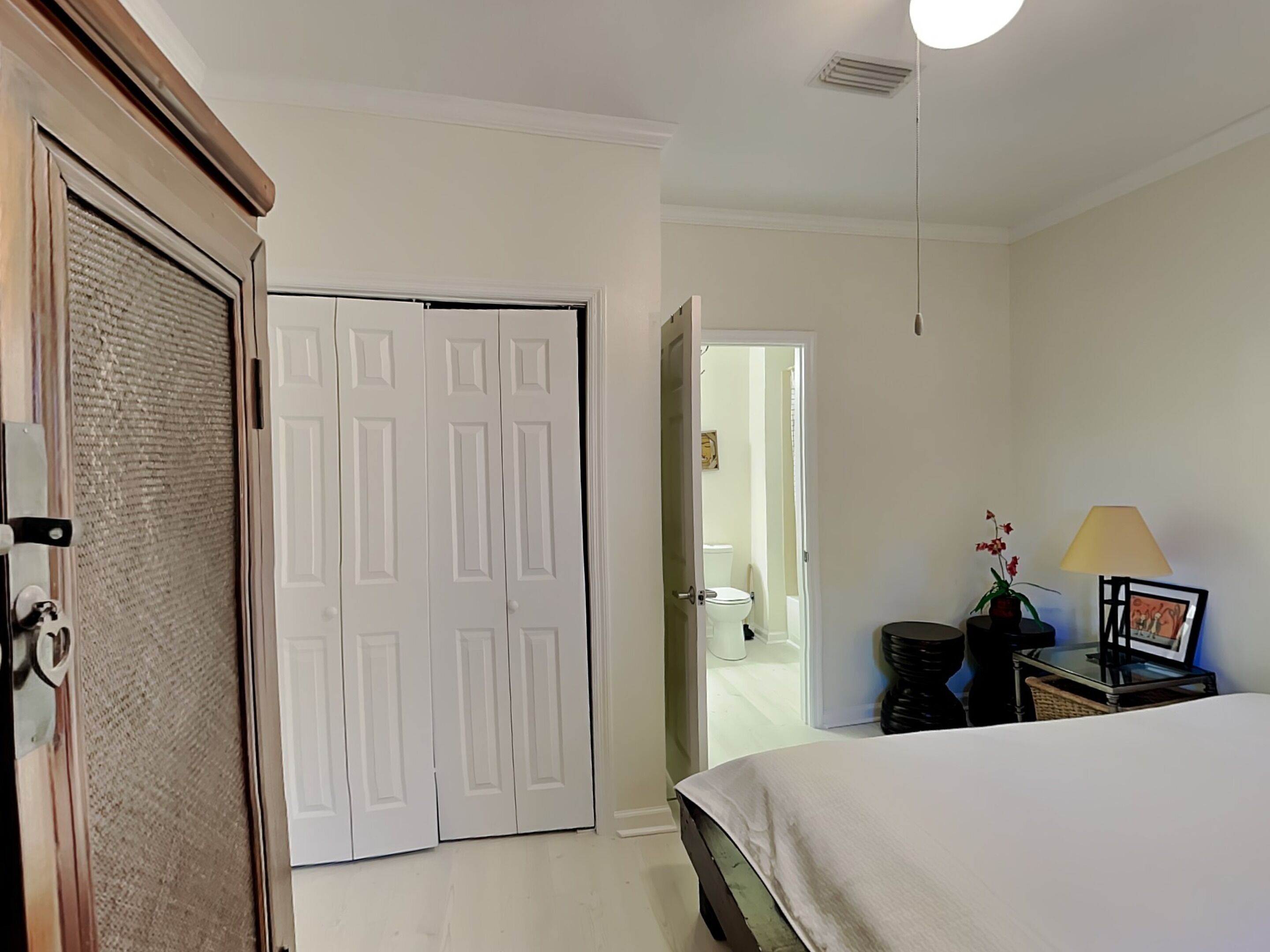 A bedroom with a bed, a wardrobe, a door leading to a bathroom, and a bedside table with a lamp and a picture frame.