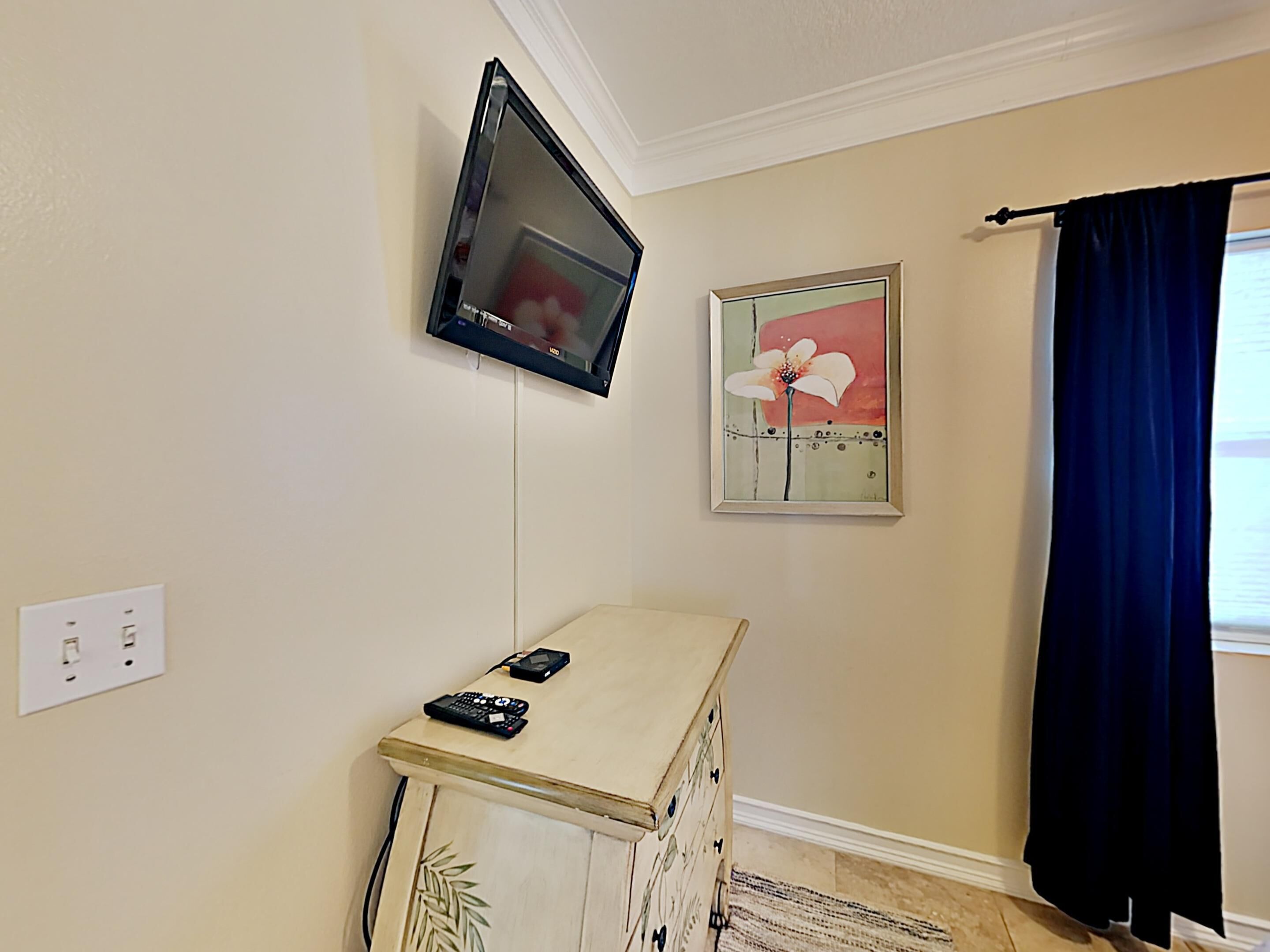 A room with a mounted flat-screen TV, a small cabinet with remote controls, a framed floral artwork, and a dark curtain.