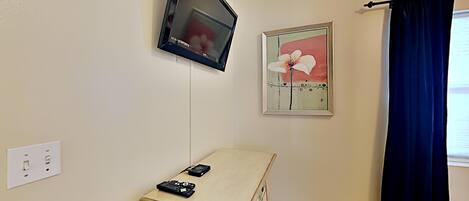 A room with a mounted flat-screen TV, a small cabinet with remote controls, a framed floral artwork, and a dark curtain.