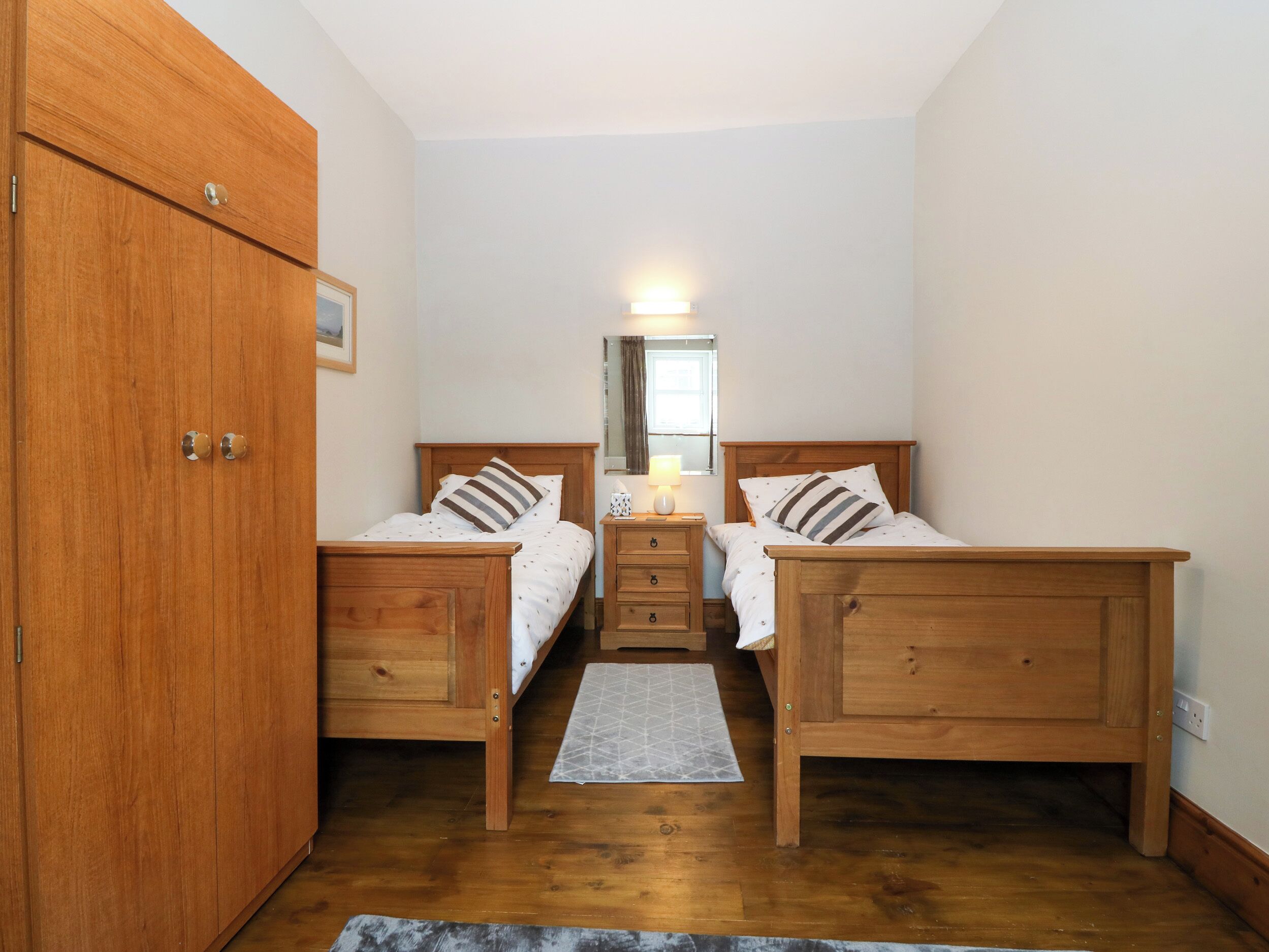 A bedroom with two wooden beds, a wooden wardrobe, a bedside table with a lamp, and a mirror on the wall.