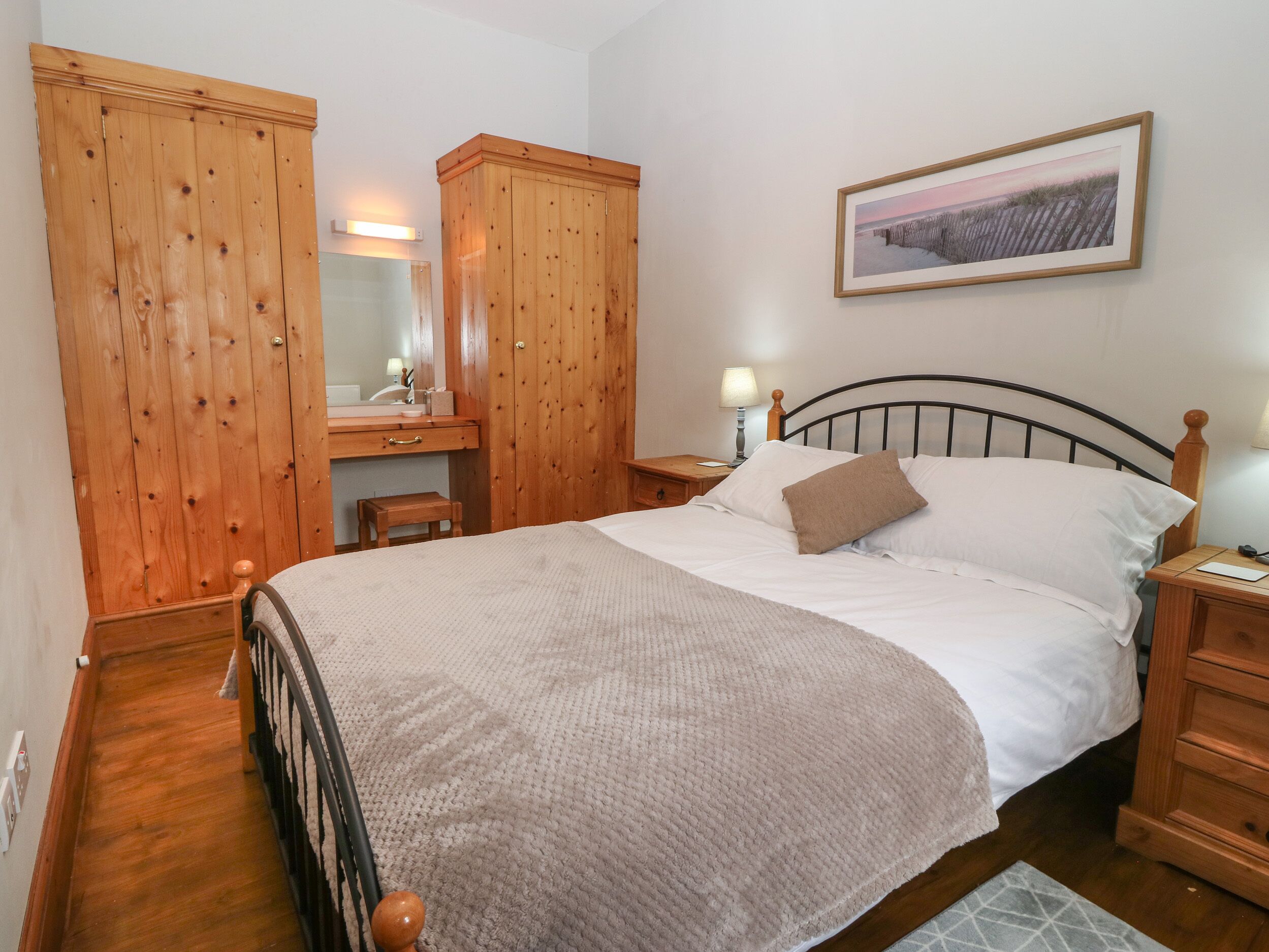 A bedroom with a bed, wooden wardrobe, a desk with a mirror, and a nightstand with a lamp.