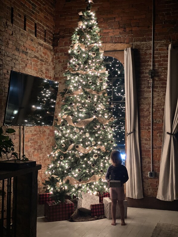 Un árbol de Navidad en una habitación con paredes de ladrillo, un televisor de pantalla plana y cortinas.