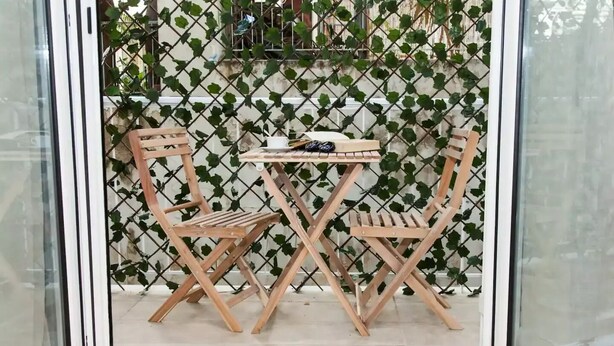 Une table et des chaises en bois sont disposées dans un espace délimité par une porte en verre, avec une clôture en treillis et de la verdure en arrière-plan.