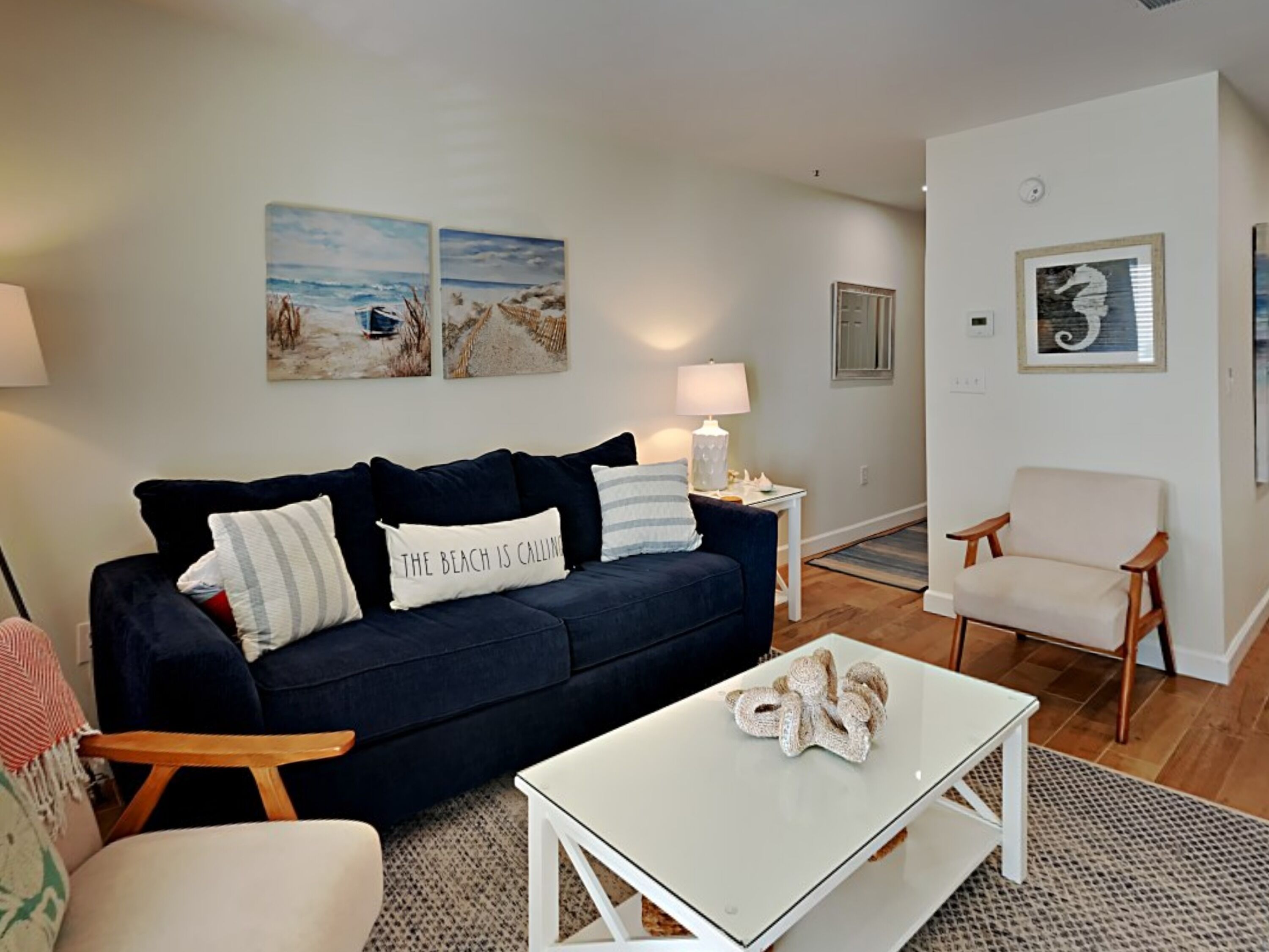 A living room with a dark blue sofa, a white coffee table, and a wooden chair. There are two wall-mounted lamps and a framed picture on the wall.