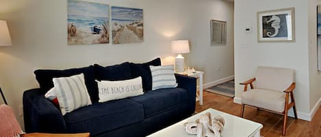 Un salon avec un canapé bleu foncé, une table basse blanche et une chaise en bois. Deux lampes sont fixées au mur, et un tableau encadré y est accroché.