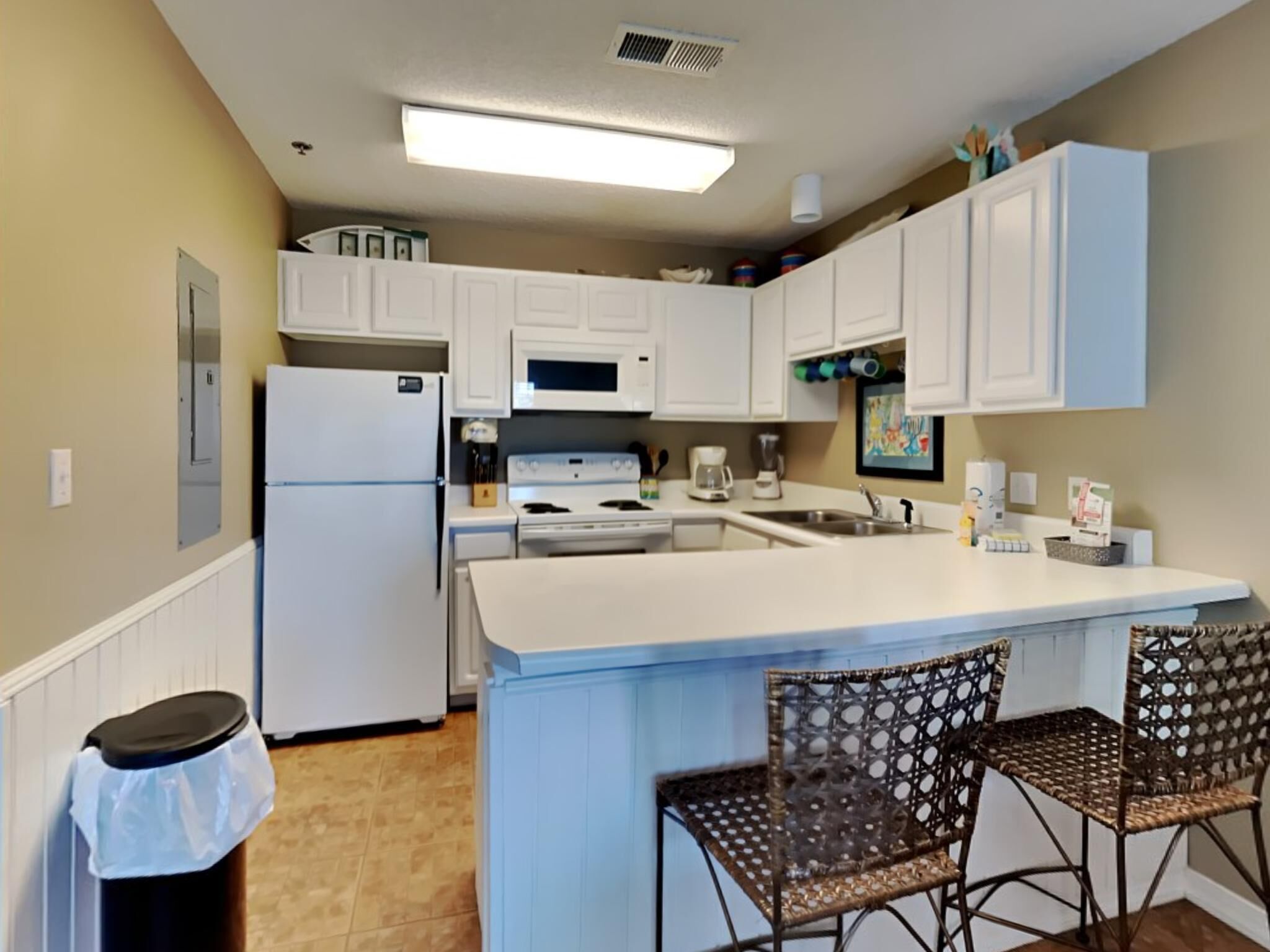 A kitchen with white cabinets, a refrigerator, a stove, and a sink. There are two wicker chairs and a trash can.