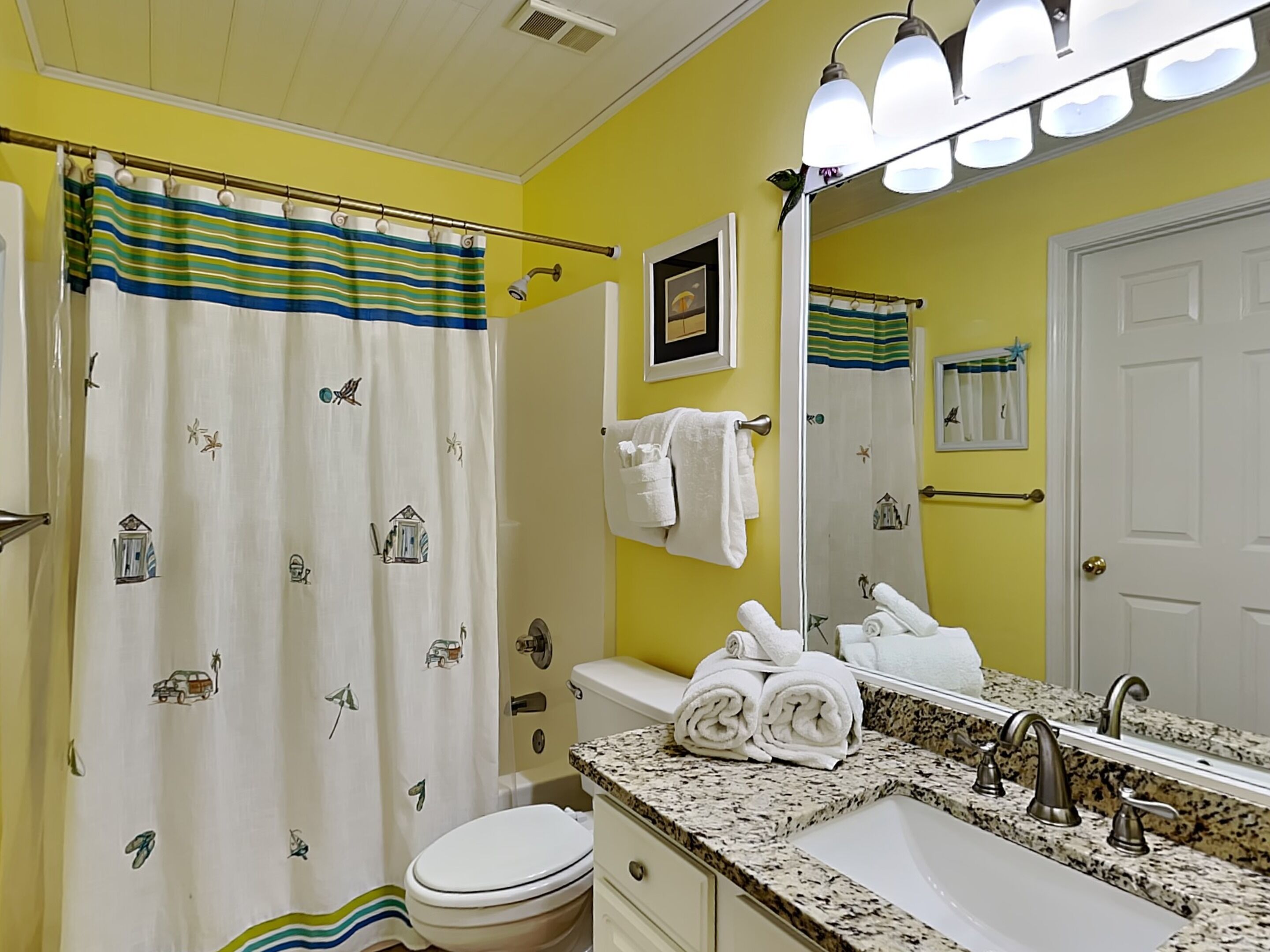 A bathroom with a yellow wall, a white toilet, a sink with a granite countertop, and a shower curtain with a striped pattern.
