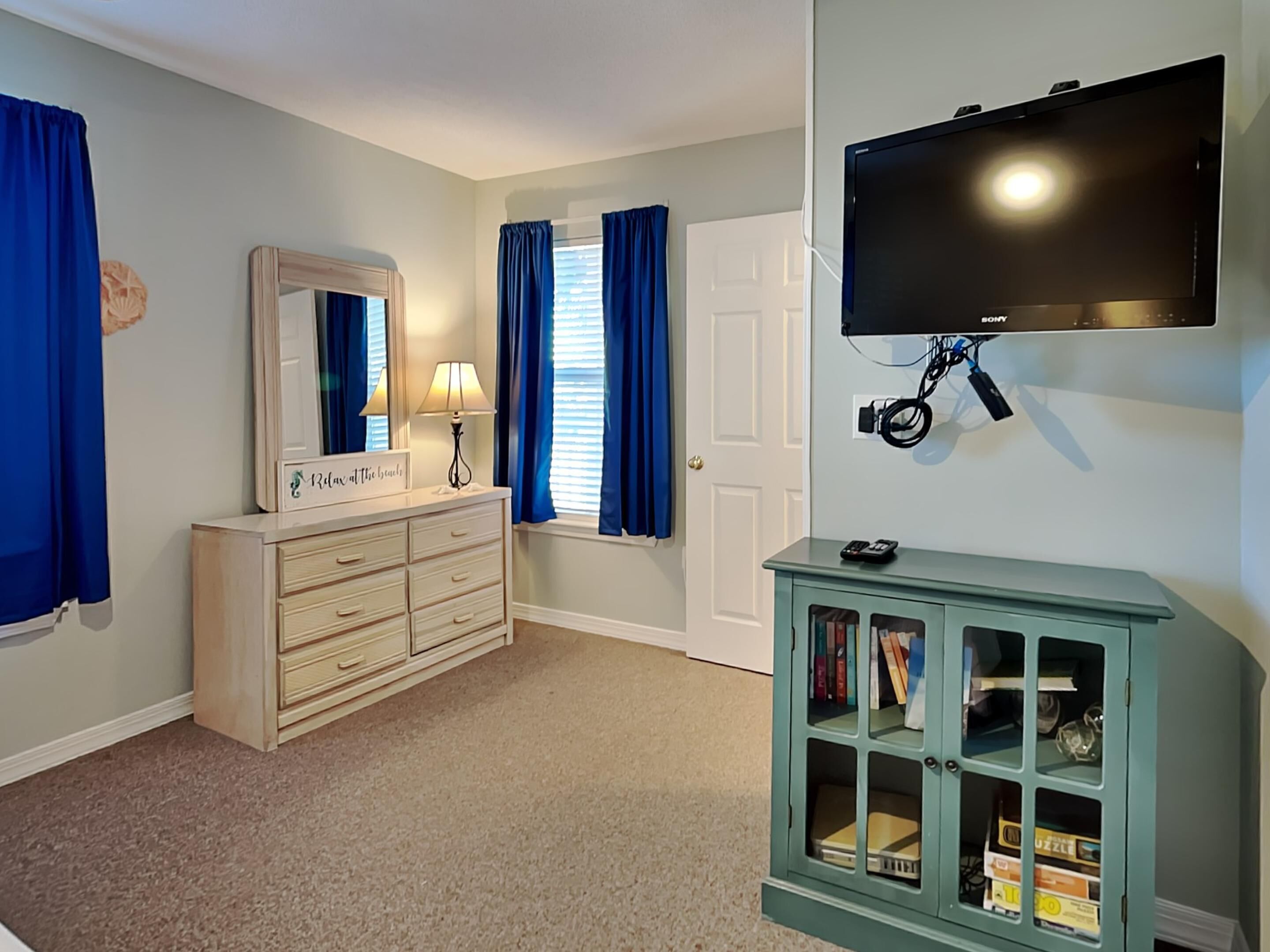 A bedroom with a dresser, a vanity with a mirror, a bookshelf, a flat-screen TV mounted on the wall, and a door.