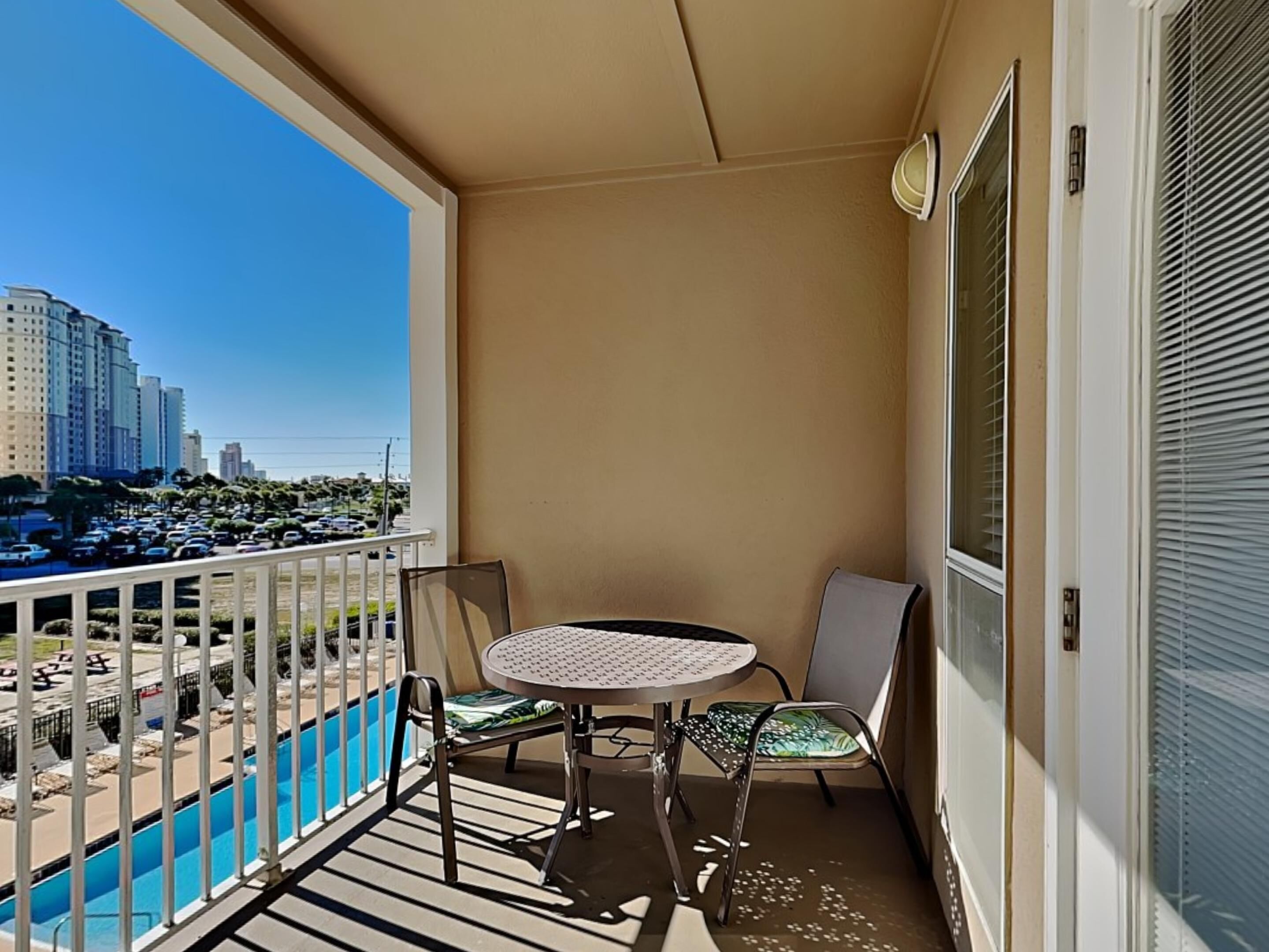 A balcony with a table and chairs, a pool, and a view of tall buildings and parked cars.