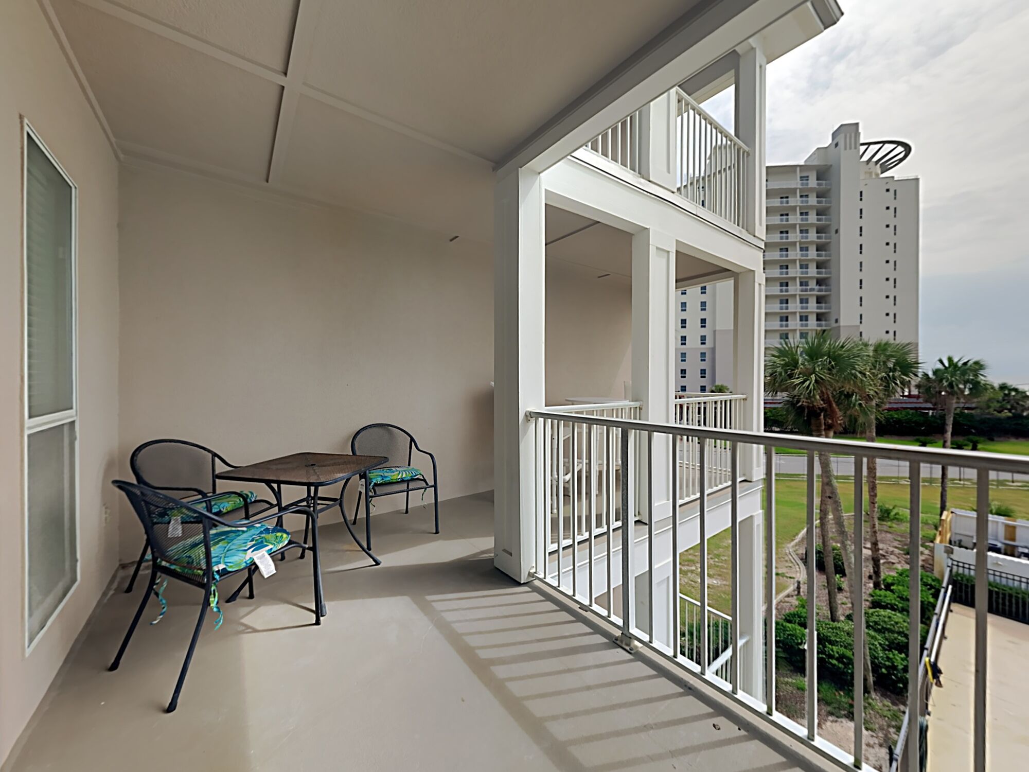 Un balcon avec une table et des chaises, une vue sur un immeuble de grande hauteur et un espace verdoyant agrémenté de palmiers.