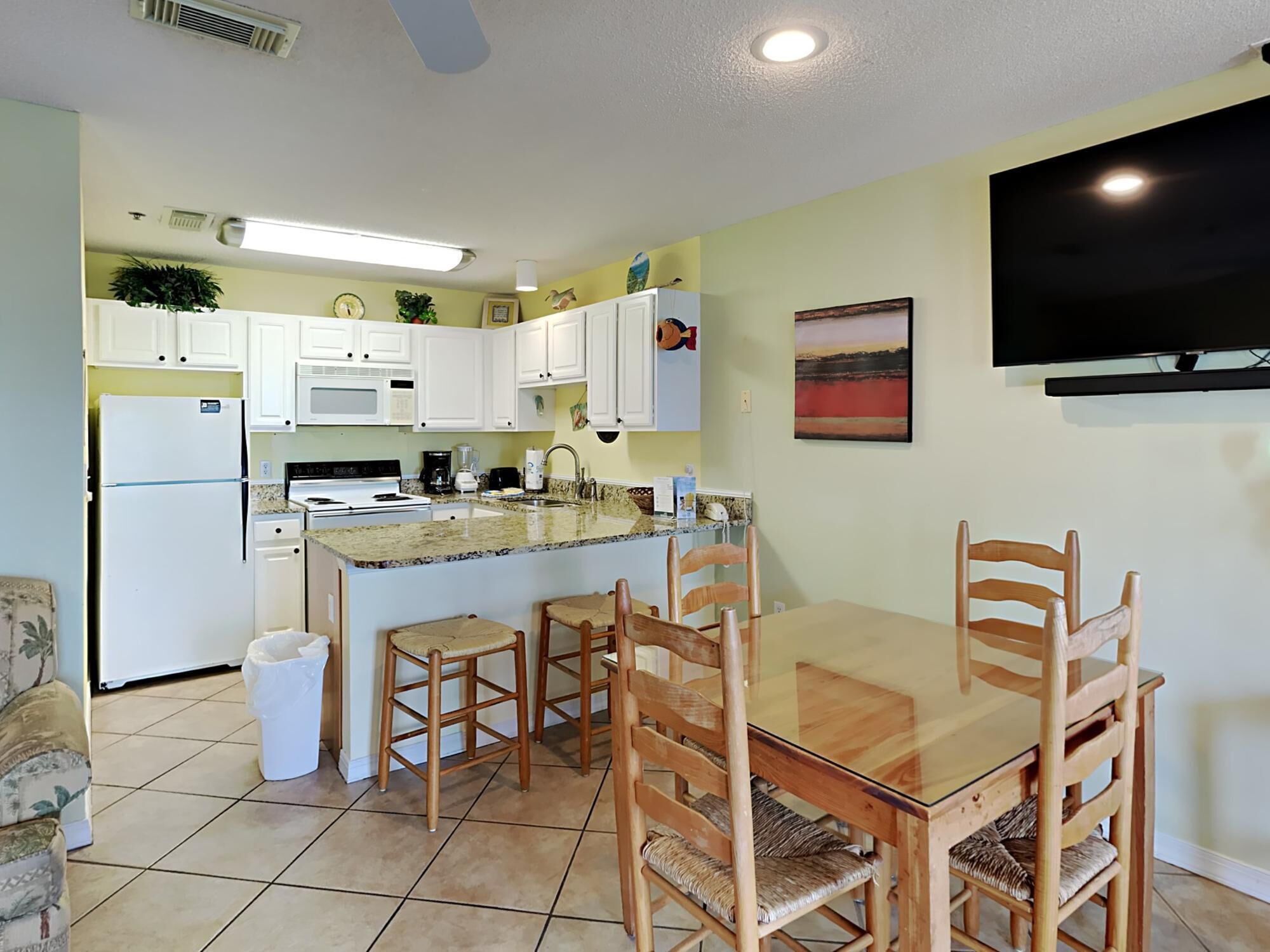 Una cocina compacta con armarios blancos, encimera de granito y una zona de comedor con sillas de madera y una mesa de vidrio.